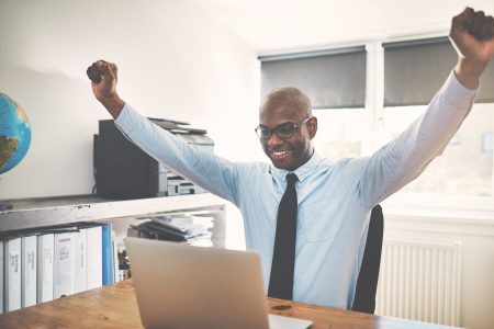 Smiling African businessman celebrating with his arms raised in the air while sitting at a desk in his home office working on a laptop