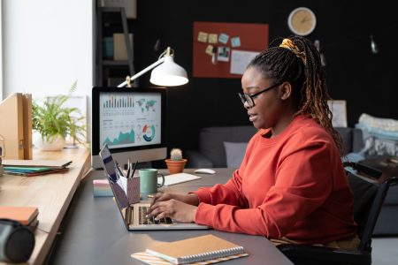 African girl in eyeglasses sitting at the table in front of the window and typing on laptop, she working online at home