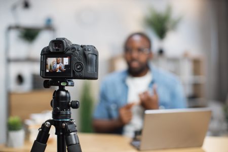 Blur background of african guy in casual wear recording new video blog while sitting at home workplace. Focus on digital camera fixed in tripod. Concept of people and social networks.