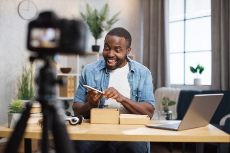 Surprised african man filming video blog at home while unpacking new modern smartphone. Concept of blogging, parcel and technology.