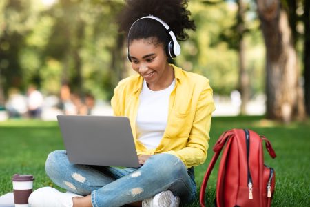 Joyful African Student Girl Learning On Laptop Watching Video Lecture Wearing Wireless Earphones Sitting In Park Outside. Modern Education, Distance Study And E-Learning Concept