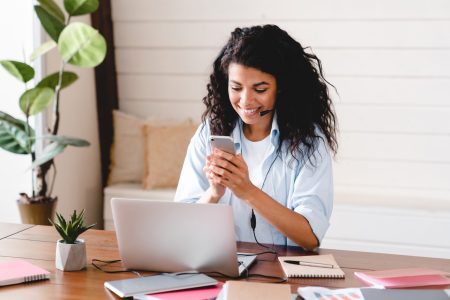 Cheerful young african girl operator IT support at workplace. Smiling young african american teen girl wear headphones video calling on laptop. Happy mixed race pretty woman student looking at computer screen watching webinar or doing video chat by webcam.